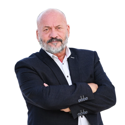 Starší muž s krátkými šedivými vlasy a plnovousem stojí před bílým pozadím v tmavém saku a bílé košili, má zkřížené ruce a neutrální výraz.