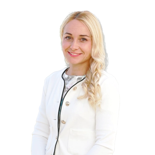 Žena se světlými vlnitými vlasy stojí proti bílému pozadí, usmívá se a má na sobě bílé sako se zdobeným lemováním a knoflíky.