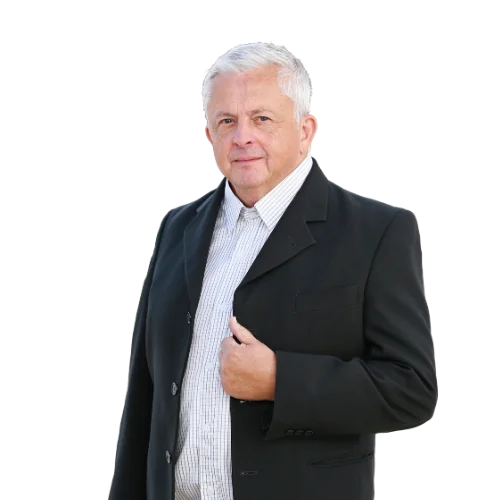 Starší muž se světlými krátkými vlasy stojí proti bílému pozadí, má na sobě černé sako a košili se vzorem. Jednou rukou se dotýká saka a působí klidně.