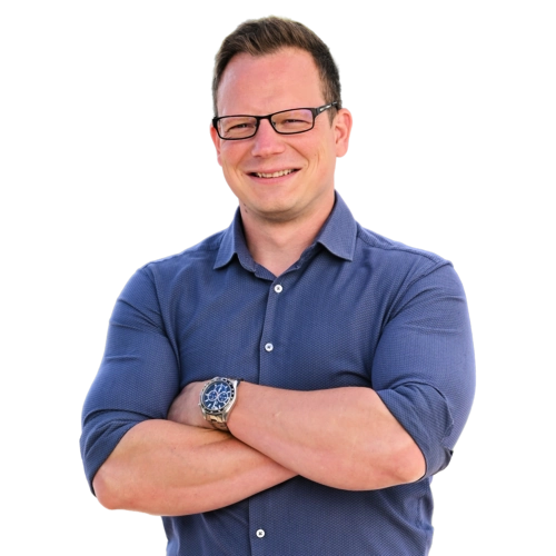 Muž s krátkými vlasy a brýlemi stojí proti bílému pozadí, má na sobě tmavě modrou košili, usmívá se a má zkřížené ruce. Nosí hodinky s kovovým páskem.