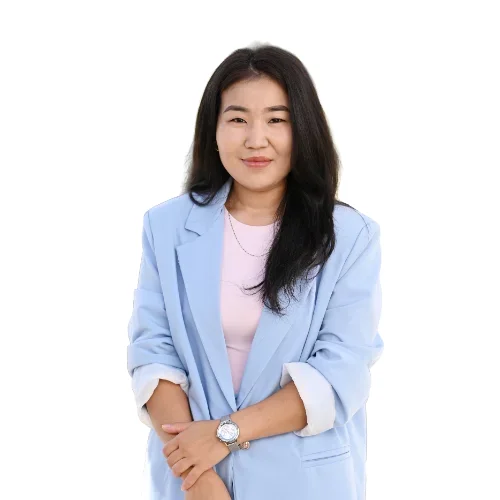 Žena s dlouhými černými vlasy stojí proti bílému pozadí, má na sobě světle modré sako, růžové tričko a hodinky. Usmívá se a má ruce volně položené před sebou.