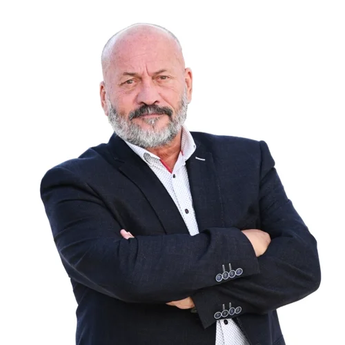 Starší muž s krátkými šedivými vlasy a plnovousem stojí před bílým pozadím v tmavém saku a bílé košili, má zkřížené ruce a neutrální výraz.